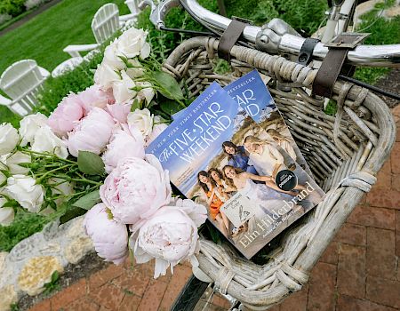 A wicker bicycle basket holds pink and white peonies alongside a book with “Five-Star” on the cover in a garden courtyard.
