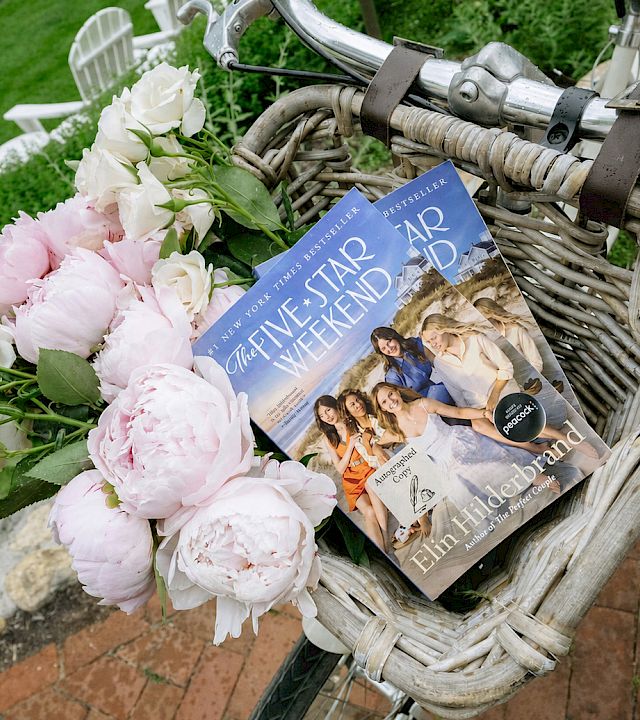 A wicker bicycle basket holds pink and white peonies alongside a book with “Five-Star” on the cover in a garden courtyard.