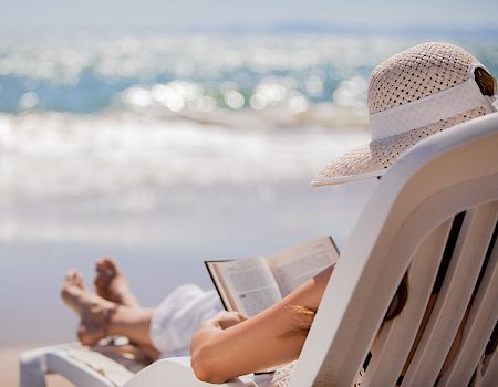 A person lounging on a beach chair by the ocean, reading a book, wearing a sunhat, with waves and sunlight in the background. End.