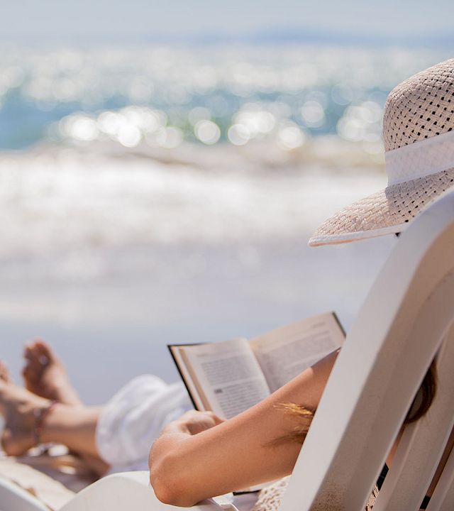 A person lounging on a beach chair by the ocean, reading a book, wearing a sunhat, with waves and sunlight in the background. End.