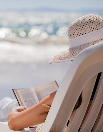 A person lounging on a beach chair reads a book by the sea, wearing a sunhat, with waves and sunlight in the background.