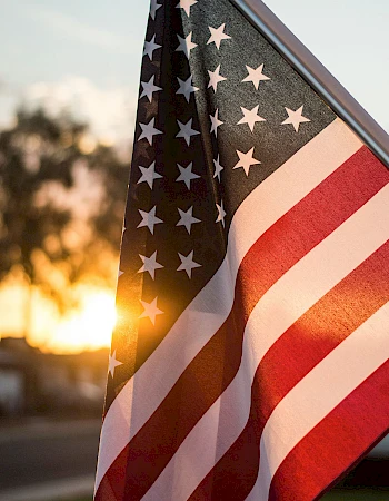A waving American flag bathed in sunset light, stars and stripes fluttering gently as the sun glows orange on the horizon.