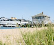 A ferry ship is docked by a seaside house while people swim near the shore under a clear blue sky.