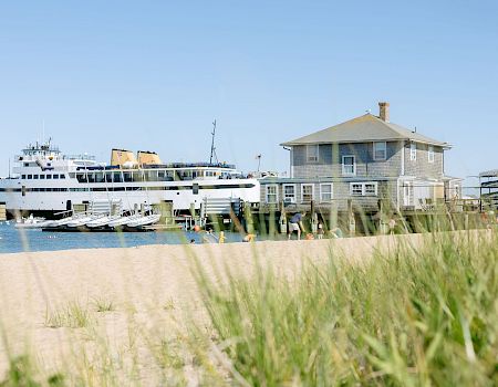 A ferry ship is docked by a seaside house while people swim near the shore under a clear blue sky.