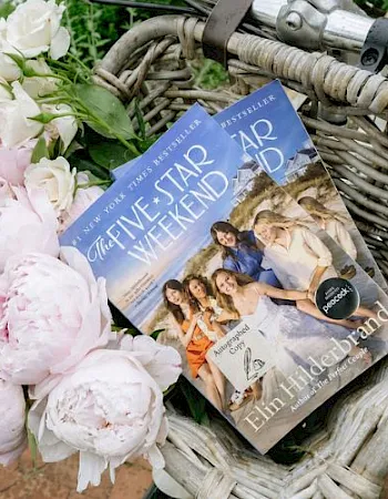 A wicker basket filled with pink and white flowers and a fashion magazine on the cover, resting on a lawn patio.
