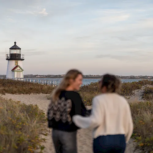 A white lighthouse with red roof stands by the coast as two people walk along a sandy path in the foreground, blurred.