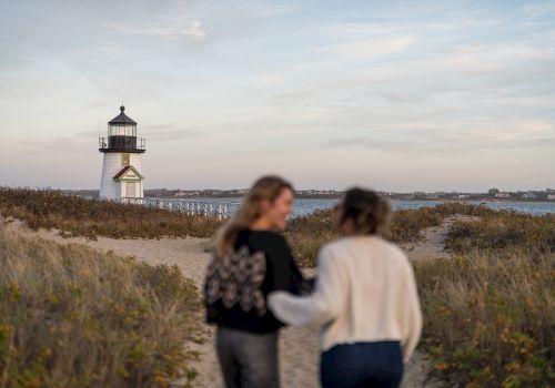 A white lighthouse with red roof stands by the coast as two people walk along a sandy path in the foreground, blurred.