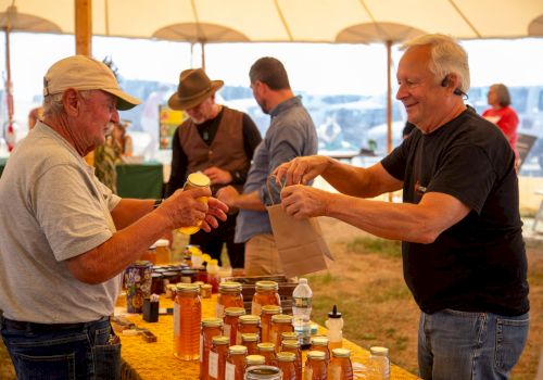 Two men under a tent pour honey and hand out samples from jars at an outdoor market table.