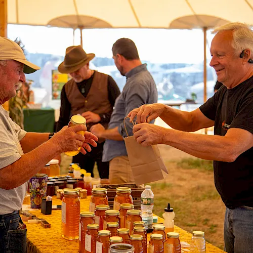 Two men under a tent pour honey and hand out samples from jars at an outdoor market table.