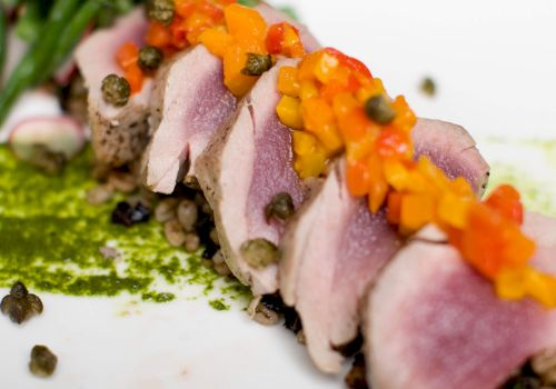 Close-up of sliced ham covered with a bright orange topping and green herbs, scattered with peppercorns on a white surface.