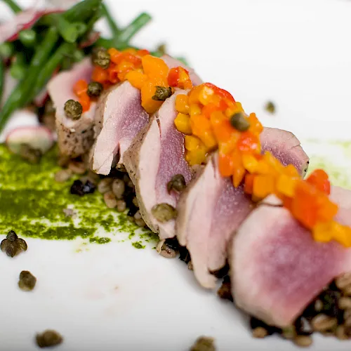 Close-up of sliced ham covered with a bright orange topping and green herbs, scattered with peppercorns on a white surface.