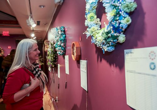 A woman in a hallway smiles at colorful hanging wreaths and ornaments on the walls during a festive indoor display.
