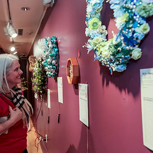 A woman in a hallway smiles at colorful hanging wreaths and ornaments on the walls during a festive indoor display.