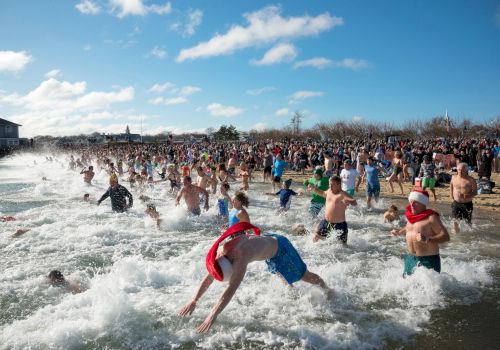 A huge crowd splashes in the surf during a cold plunge at the beach, with swimmers tossing water as waves surge.