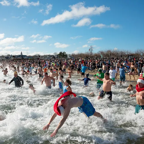 A huge crowd splashes in the surf during a cold plunge at the beach, with swimmers tossing water as waves surge.