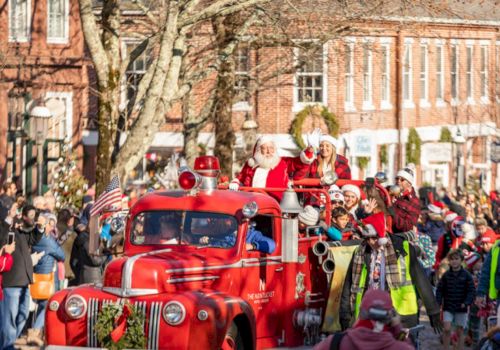 Santa and elves ride a bright red fire truck in a street Christmas parade while crowds cheer in festive winter outfits.