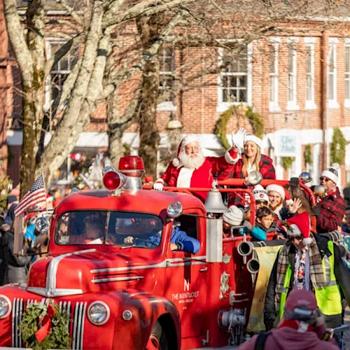 Santa and elves ride a bright red fire truck in a street Christmas parade while crowds cheer in festive winter outfits.