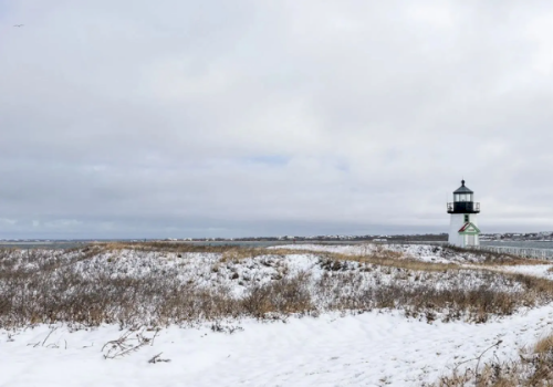 Snow-covered coastal dunes with a small lighthouse and an overcast sky.