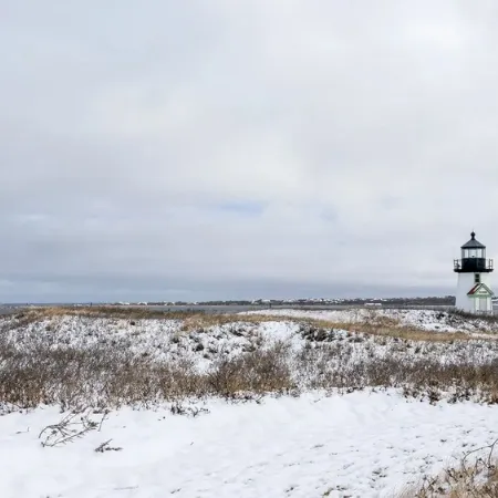 Snow-covered coastal dunes with a small lighthouse and an overcast sky.