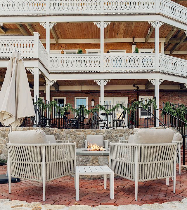 Outdoor patio with white railings, hanging plants, brick fire pit, and beige cushioned chairs beneath a covered wooden pavilion.