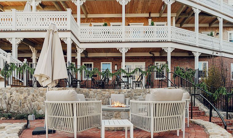 Outdoor patio with white railings, hanging plants, brick fire pit, and beige cushioned chairs beneath a covered wooden pavilion.