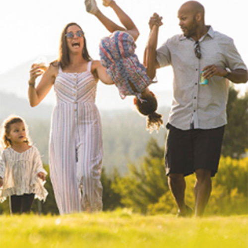 A family of four enjoys a sunny day outdoors, with the father playfully swinging a child. The mother and another child walk beside them.