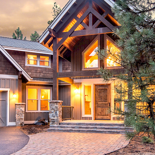 A beautiful, large modern home with multiple peaked roofs, large windows, and a driveway, surrounded by trees at dusk, with warm interior lights.