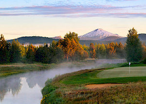 The image shows a serene golf course near a river with mist, surrounded by trees. In the background, there's a snow-capped mountain under a colorful sky.