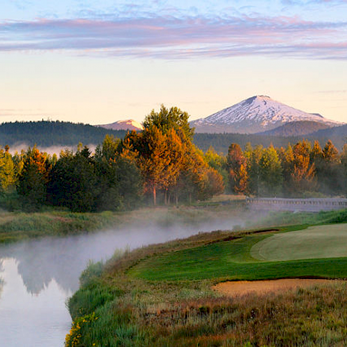 The image shows a serene golf course near a river with mist, surrounded by trees. In the background, there's a snow-capped mountain under a colorful sky.