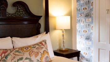 A cozy bedroom features an ornate wooden headboard, floral pillow, elegant lamp on a side table, and patterned wallpaper in the corner.