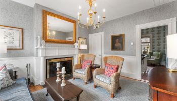 A cozy living room with patterned wallpaper, wicker chairs, a fireplace, chandelier, and art on the walls. Elegant and inviting space.