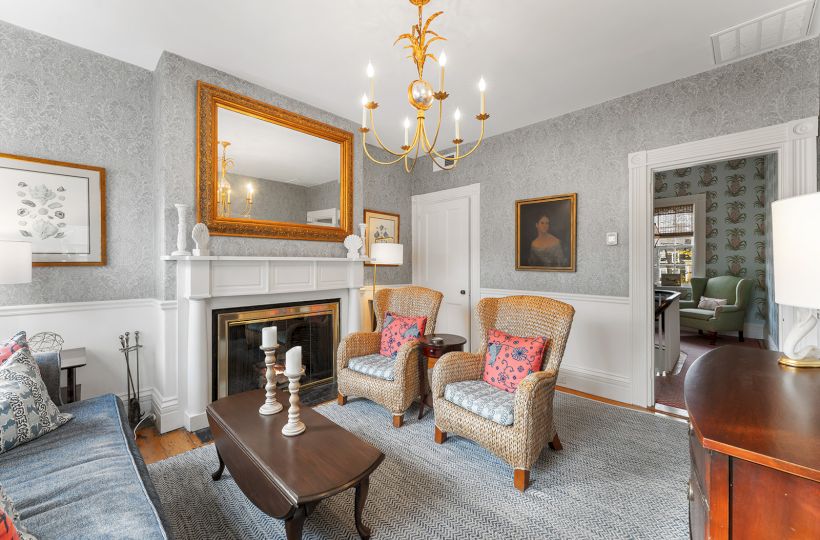 A cozy living room with patterned wallpaper, wicker chairs, a fireplace, chandelier, and art on the walls. Elegant and inviting space.